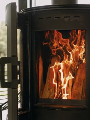 Llar de foc negra moderna amb foc cremant i llenya sobre un suport metàl·lic amb una gran finestra al fons. Moments acollidors, càlids i tranquils en temporada de fred, escalfador a la cabana